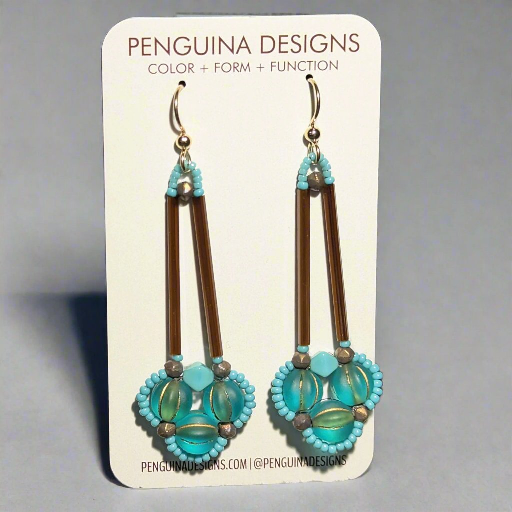 Aqua and brown earrings on a white card. The earrings have a clover of three aqua beads outlined in tiny turquoise beads. The clover shape is suspended from a pair of long, thin brown tube beads. 