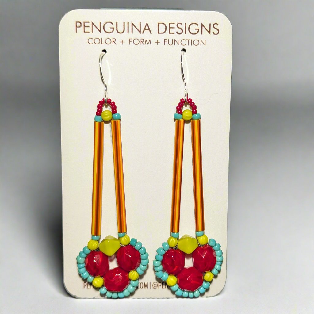 brightly colored earrings on a white card. The earrings have a clover of red beads outlined in tiny turquoise beads. The clover shape is suspended from a pair of long, thin red and orange striped tube beads. 