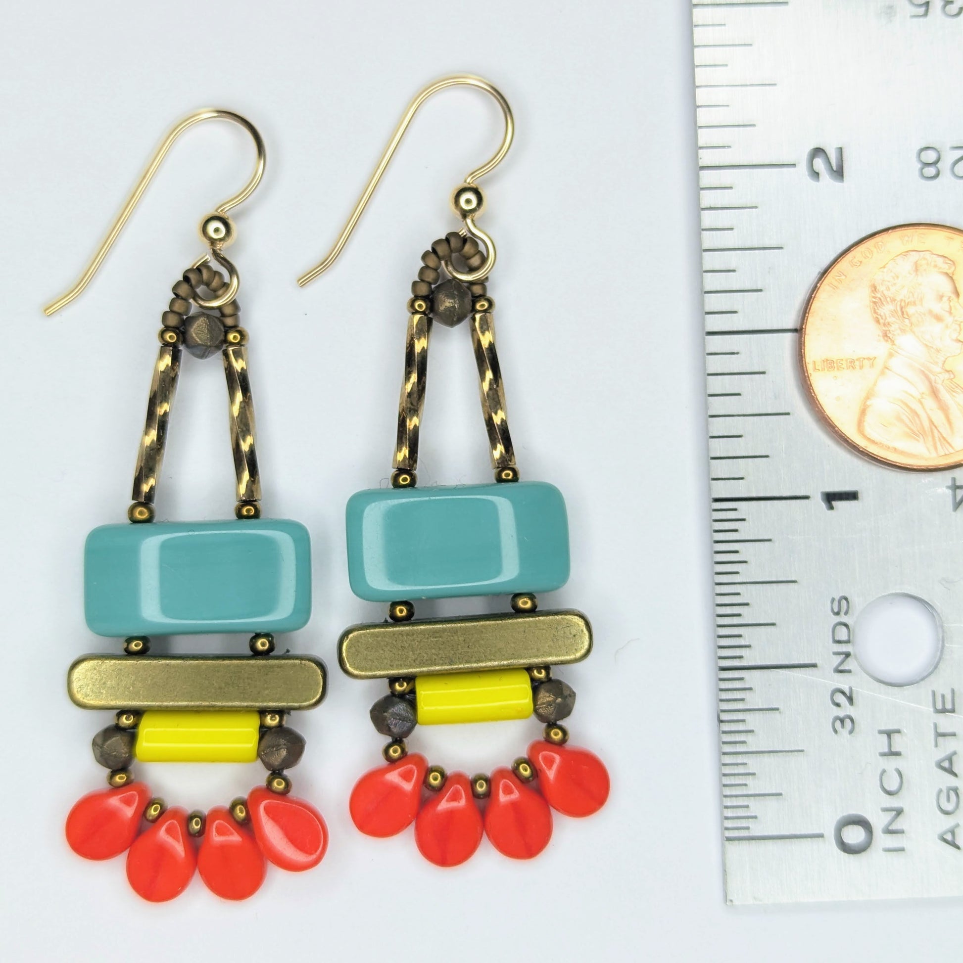 Long earrings laying next to a ruler with a penny on it. The earrings each have a wide turquoise rectangle stacked on top of a brass colored bar. Under the bar there is a yellow tube and a swag of four warm red petal beads.