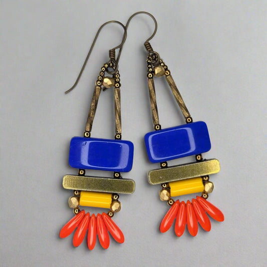 Long earrings that have a wide blue rectangle stacked on top of a brass colored bar. Under the bar there is a light orange tube and a swag of five red dagger beads.